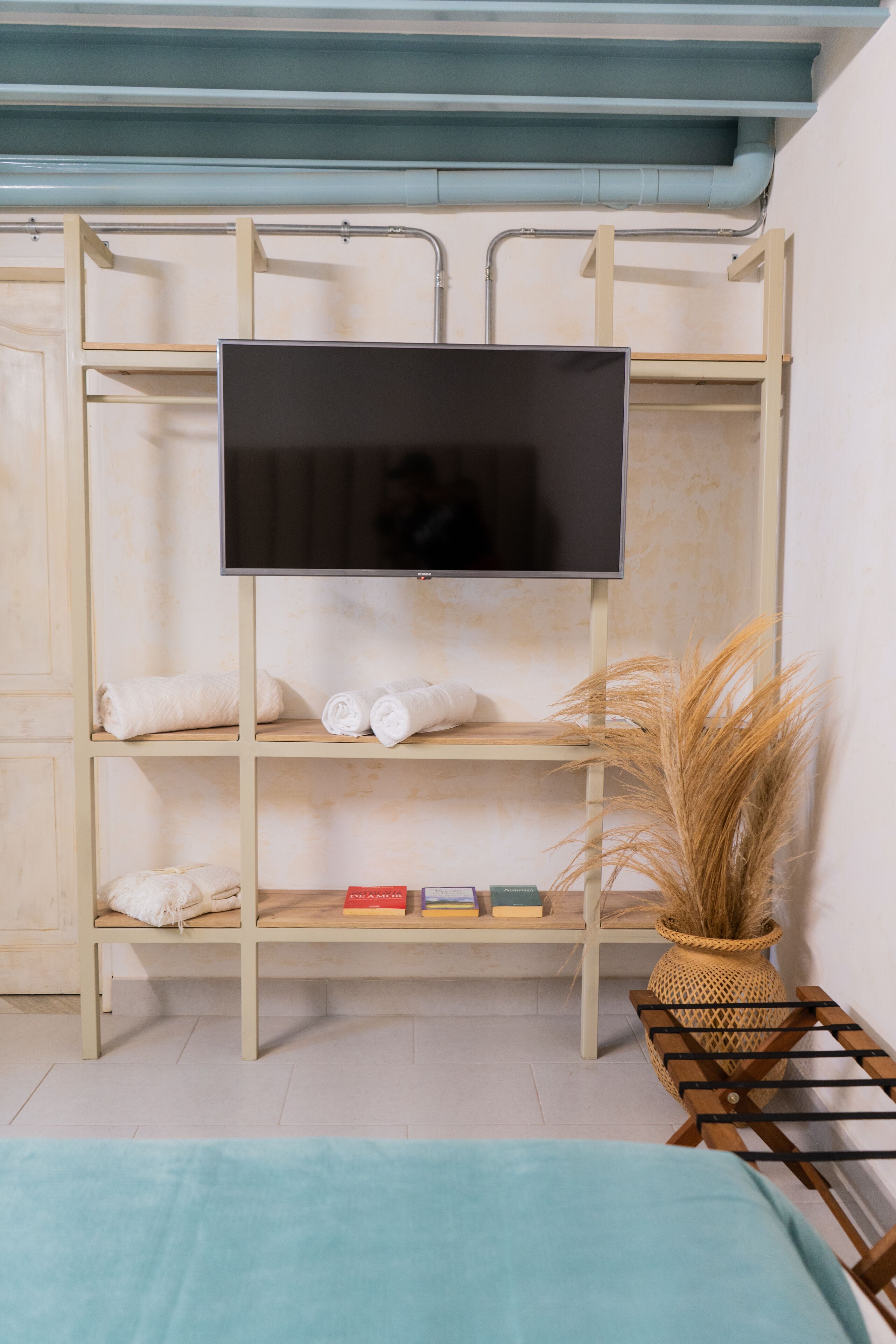 Una cama litera con un televisor de pantalla plana montado encima, un estante con toallas y libros, y una cesta tejida con plantas secas.
