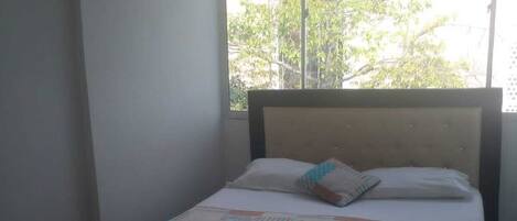 Una cama individual con cabecera, una ventana con vistas a árboles y edificios, y una mesita con una bolsa encima.