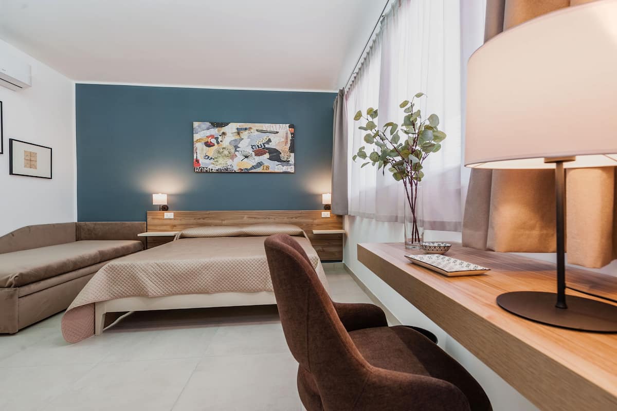 A modern hotel room with a bed, a desk with a lamp, a chair, and a wall-mounted painting.