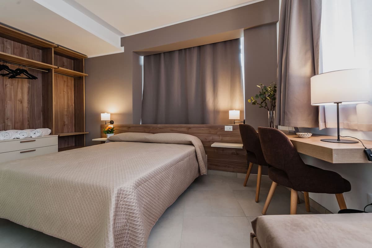 A modern hotel room with a bed, bedside tables, a desk with a chair, a lamp, and a wardrobe.