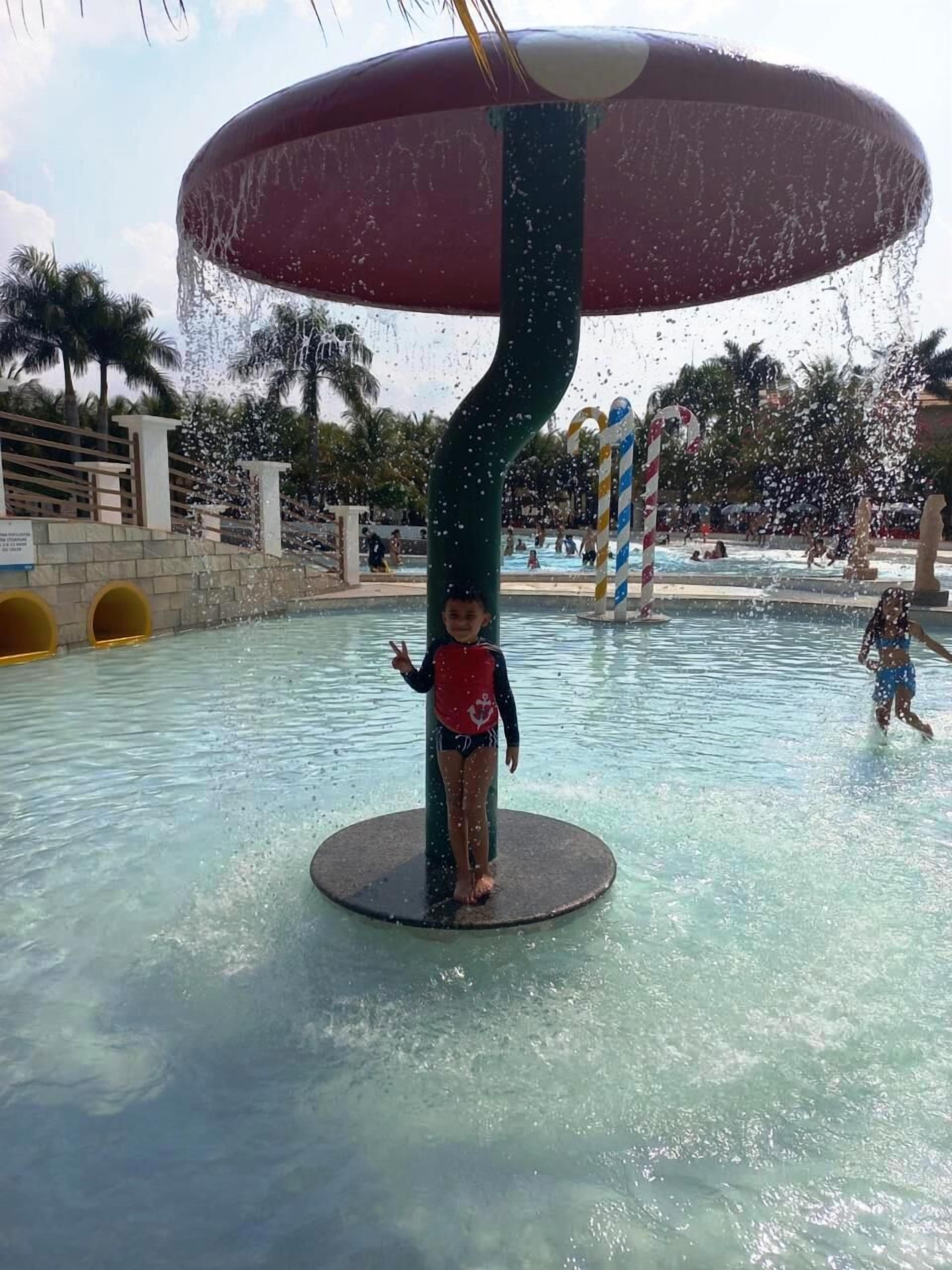 A water play area with a large umbrella structure, a pool, and palm trees in the background.