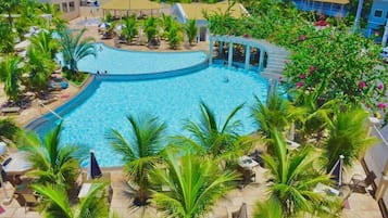 A large, clear swimming pool surrounded by palm trees and lounge chairs, with a view of hotel buildings in the background.