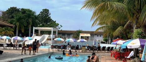 A swimming pool area with people swimming and lounging, surrounded by umbrellas and chairs.