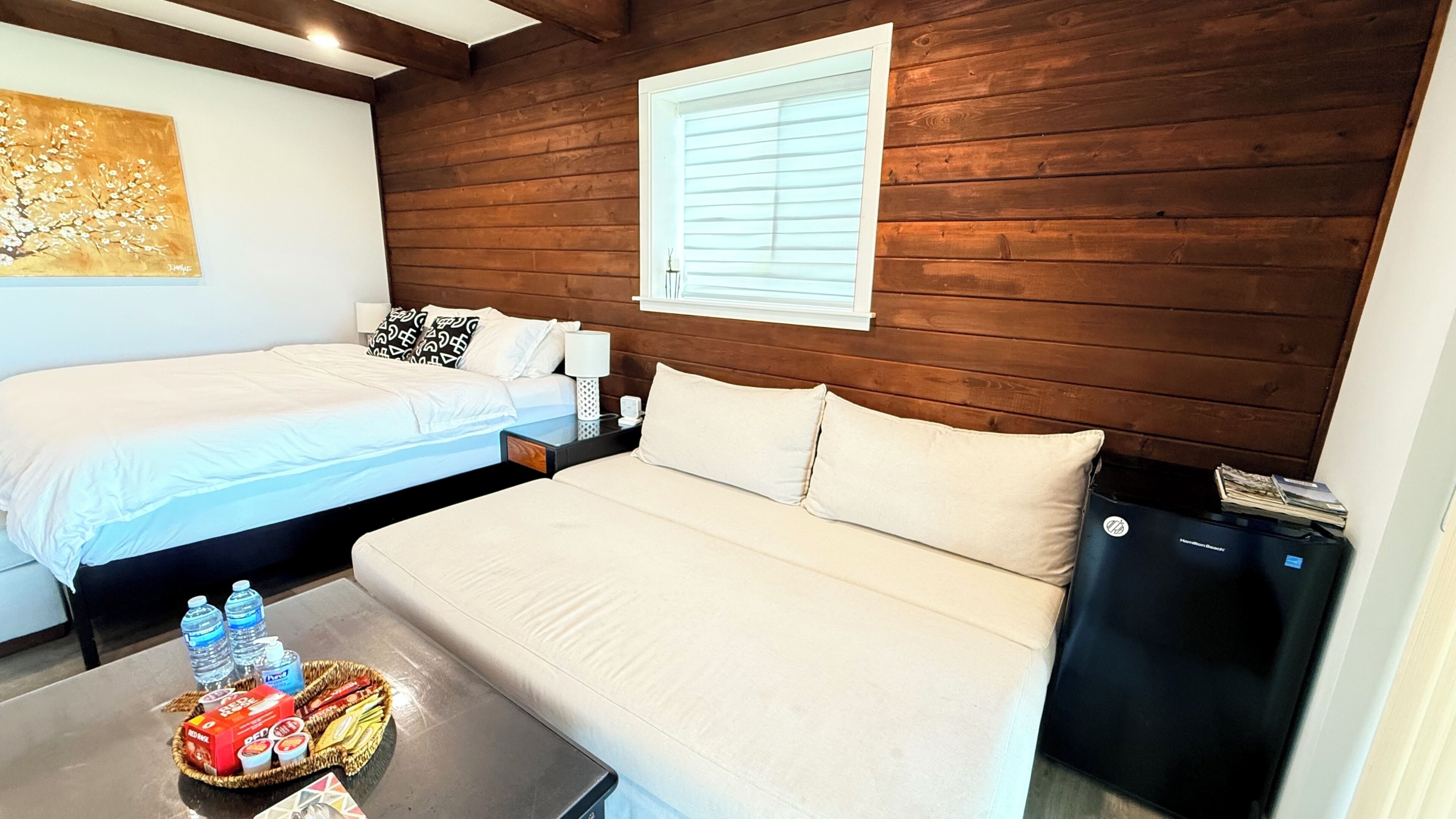 A room with two beds, a sofa, a small table with snacks, a water dispenser, and a window with blinds.