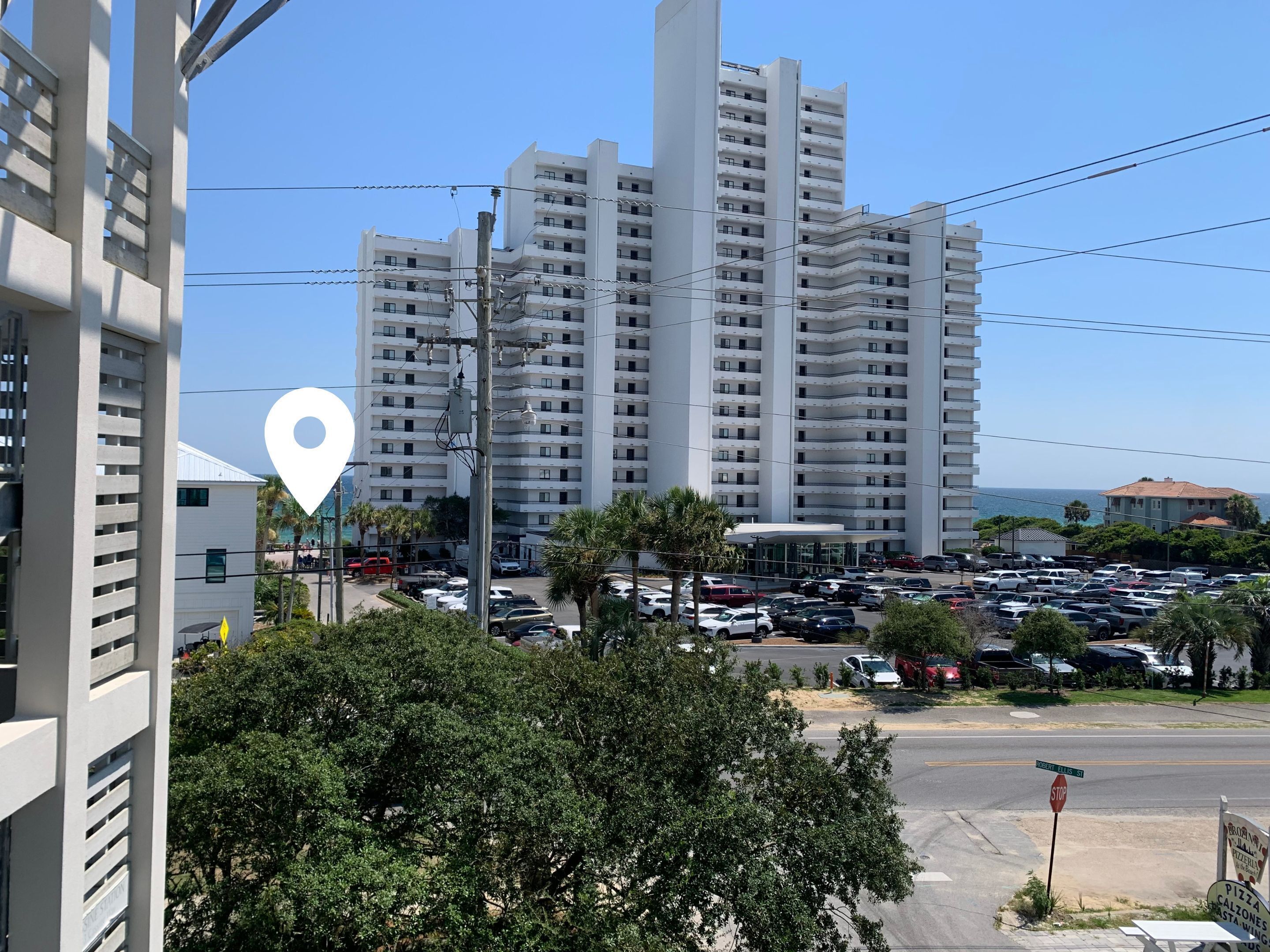 Un immeuble de grande hauteur doté de nombreux balcons, un parking où sont garées différentes voitures, et une vue dégagée sur l’océan en arrière-plan.