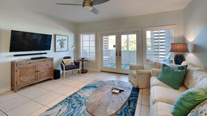 A spacious living room with a ceiling fan, a flat-screen TV, a wooden coffee table, and a large window offering a view of the outdoors. - Cassine Station #305 by Southern Vacation Rentals (Santa Rosa Beach)