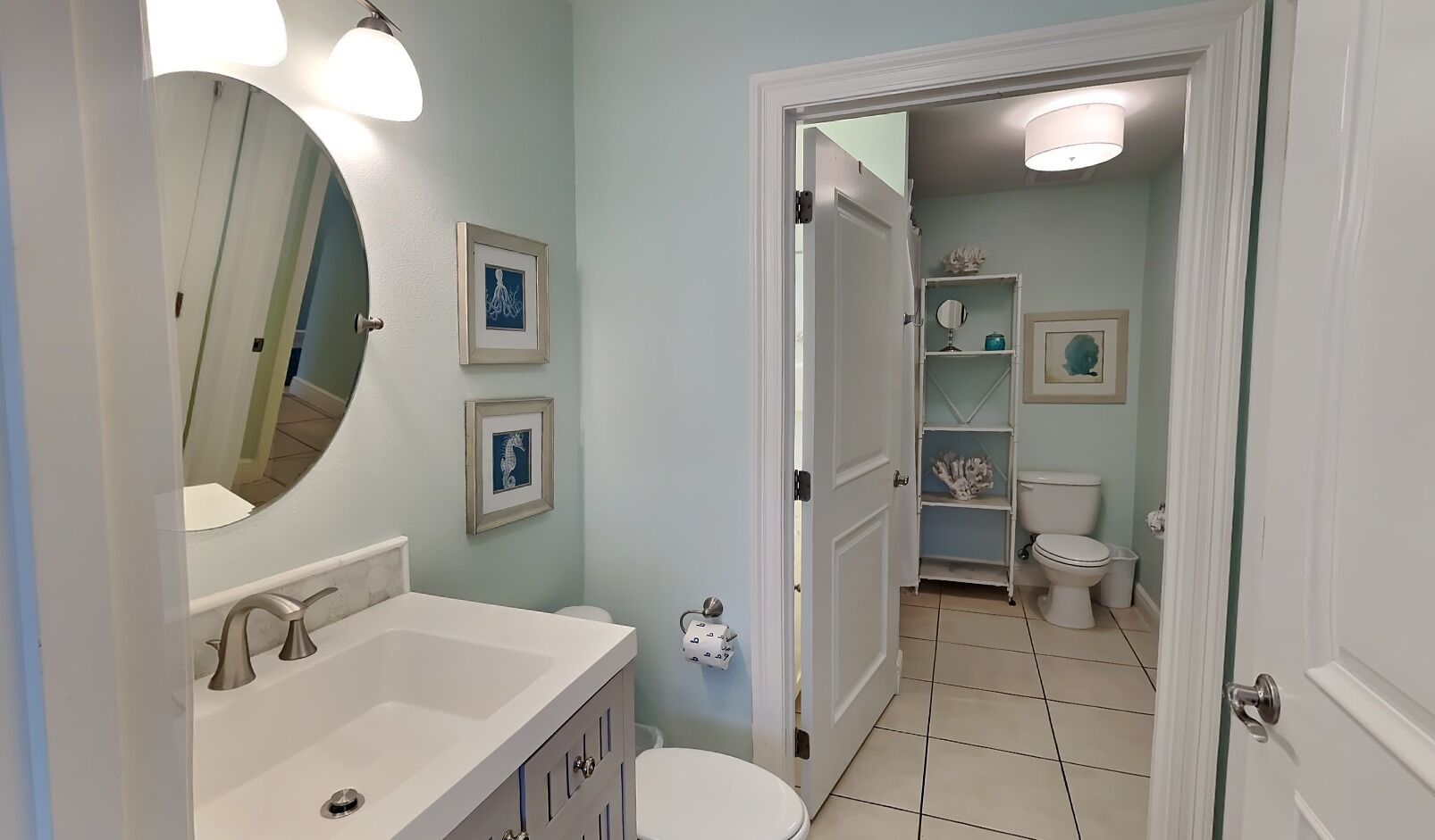 Ein Badezimmer mit einem weißen Waschbecken, einem Spiegel und gerahmten Bildern an der Wand. Eine Toilette und ein Regal mit Dekorationselementen sind ebenfalls vorhanden.