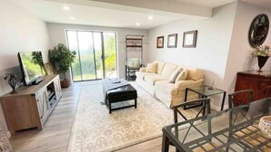 A spacious living room with a sofa, a coffee table, a TV, and a sliding glass door leading to a balcony. - 2BR Private Suite · West Vancouver · Landmark View · Prime Location (West Vancouver)