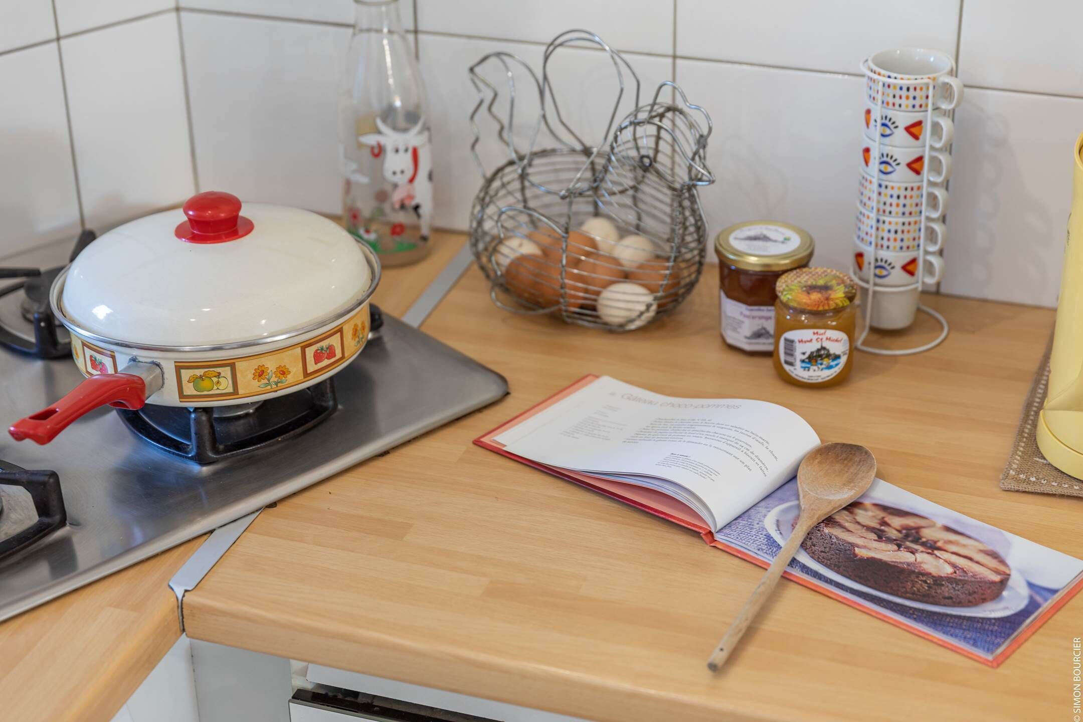 A kitchen counter with a stove, a cookbook, a wooden spoon, a jar of jam, and a basket of eggs.