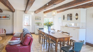 A cozy living room with a red leather sofa, wooden dining table, and chairs, a kitchen area with white cabinets and a window, and a tiled floor. - Cottage Roazhon (CHERRUEIX)