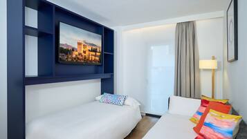 Habitación de hotel con cama, televisión montada en un estante azul, ventana con cortinas y una lámpara.