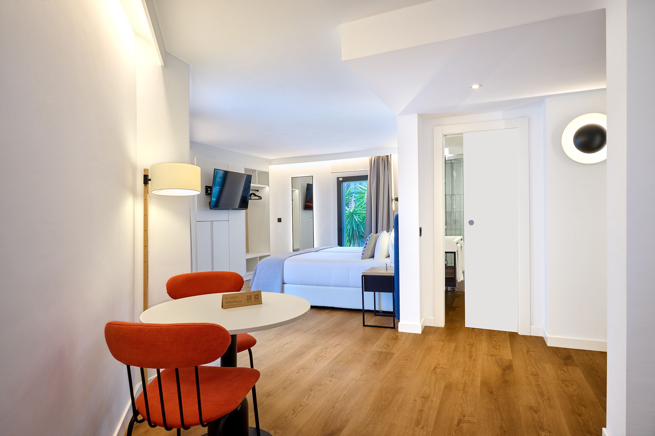 Habitació d'hotel moderna amb un llit, una taula petita amb dues cadires, una televisió penjada a la paret i un paviment de fusta.