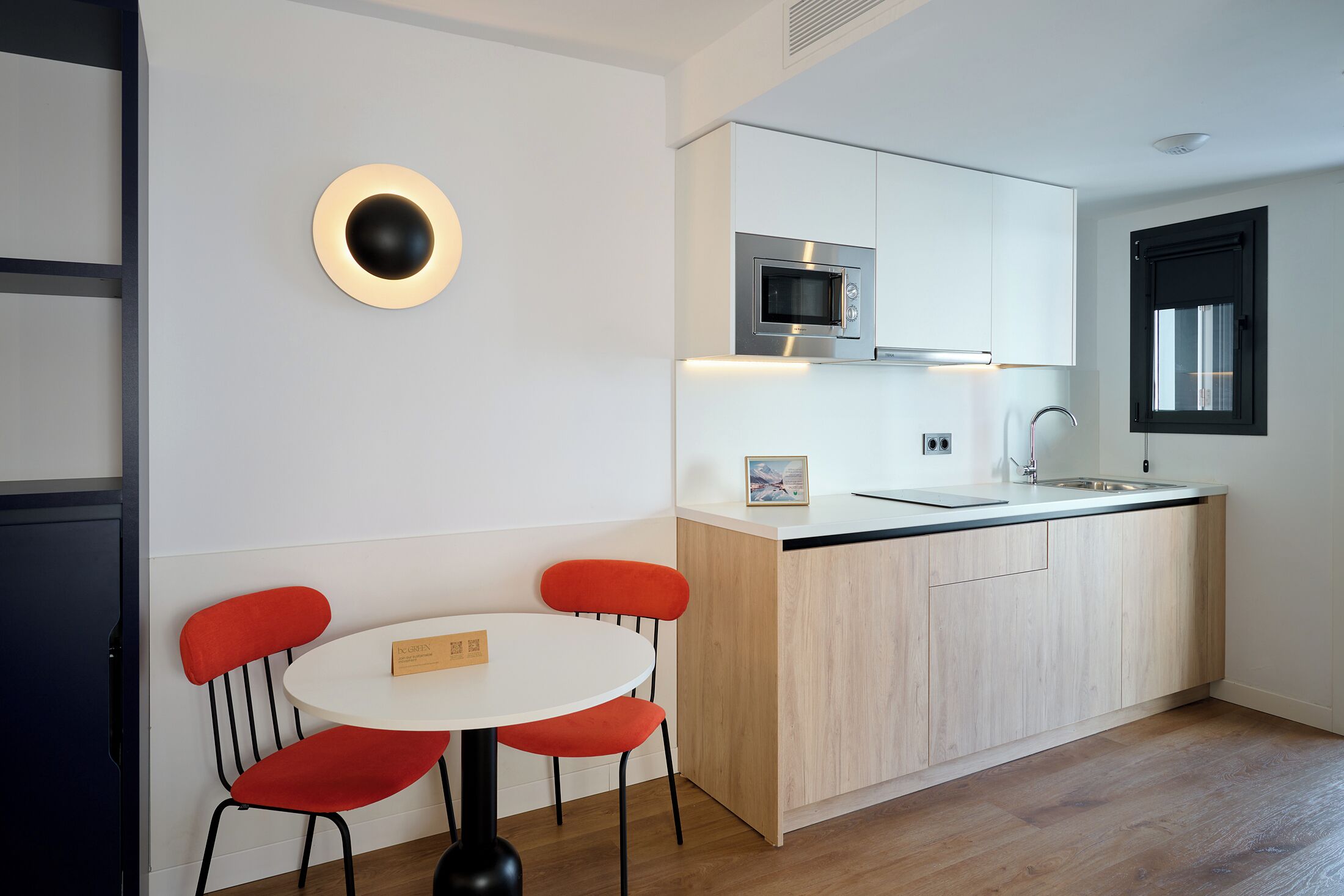 Une cuisine moderne avec un plan de travail blanc, un four à micro-ondes encastré et un évier. On y trouve une table ronde avec deux chaises rouges et une petite plaque nominative.