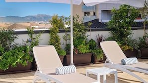 A rooftop terrace with two white lounge chairs, a small table, and a large umbrella providing shade. The terrace is surrounded by potted plants and features a view of distant mountains. - Apartamentos Luna Suites Granada (Granada)