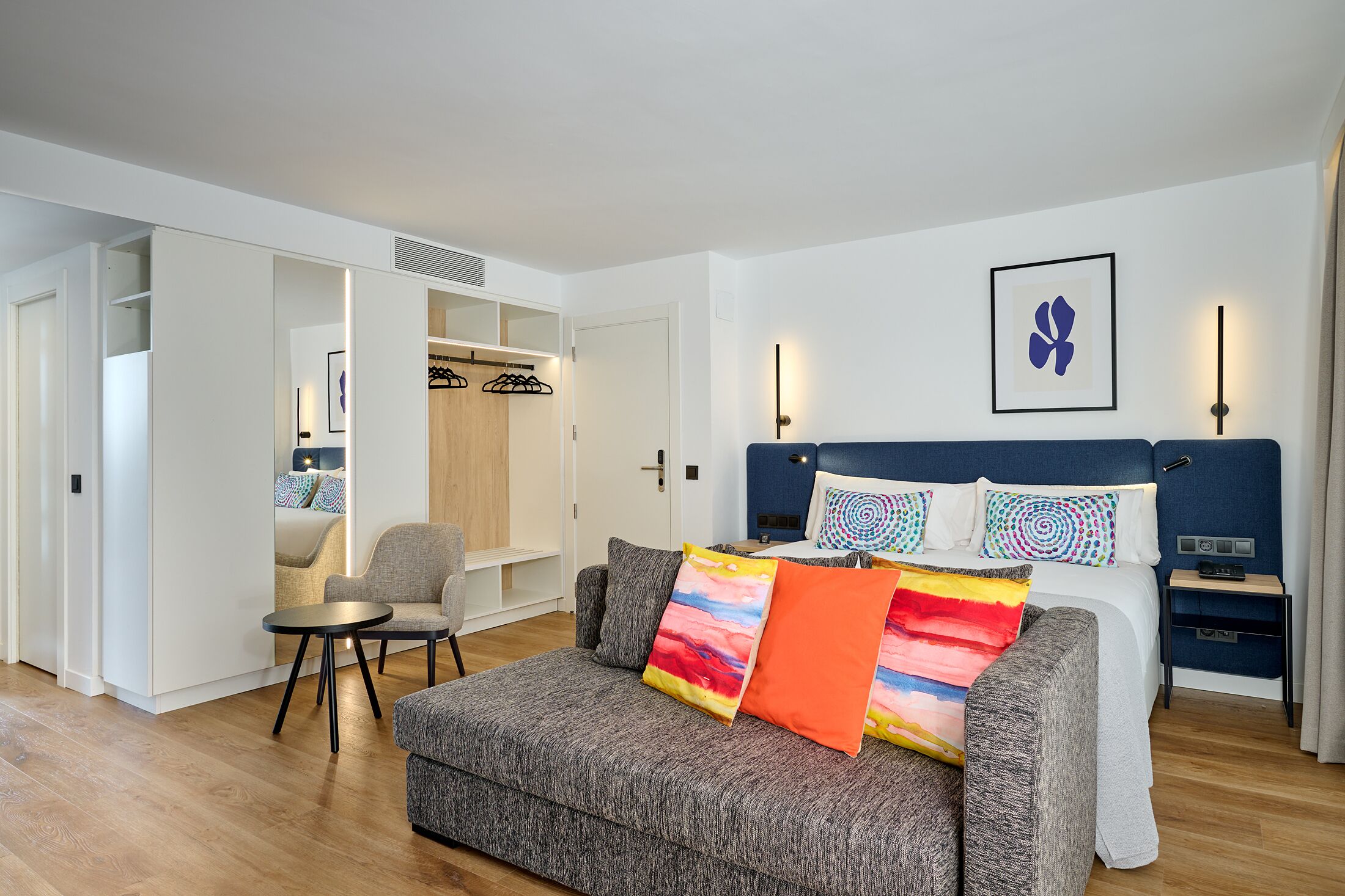Un dormitorio moderno con un sofá gris, una cama con almohadas coloridas, una mesita de noche y un armario.