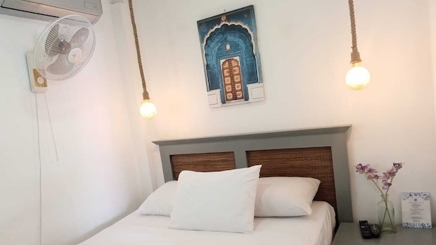 A neatly made bed with white linens, a wooden headboard, a wall-mounted air conditioner, a decorative painting above the bed, and a bedside table with a vase of flowers and a remote control. - Apartamentos Magallanes Romana (La Romana)