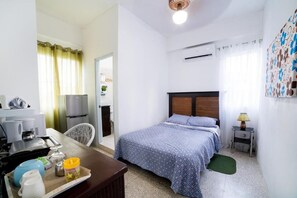 A hotel room with a bed, a desk with a computer, a chair, and a small table with a lamp.