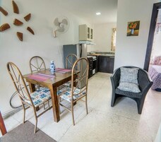 A dining area with a wooden table and chairs, a black chair with a patterned cushion, a fan, and a kitchen in the background.