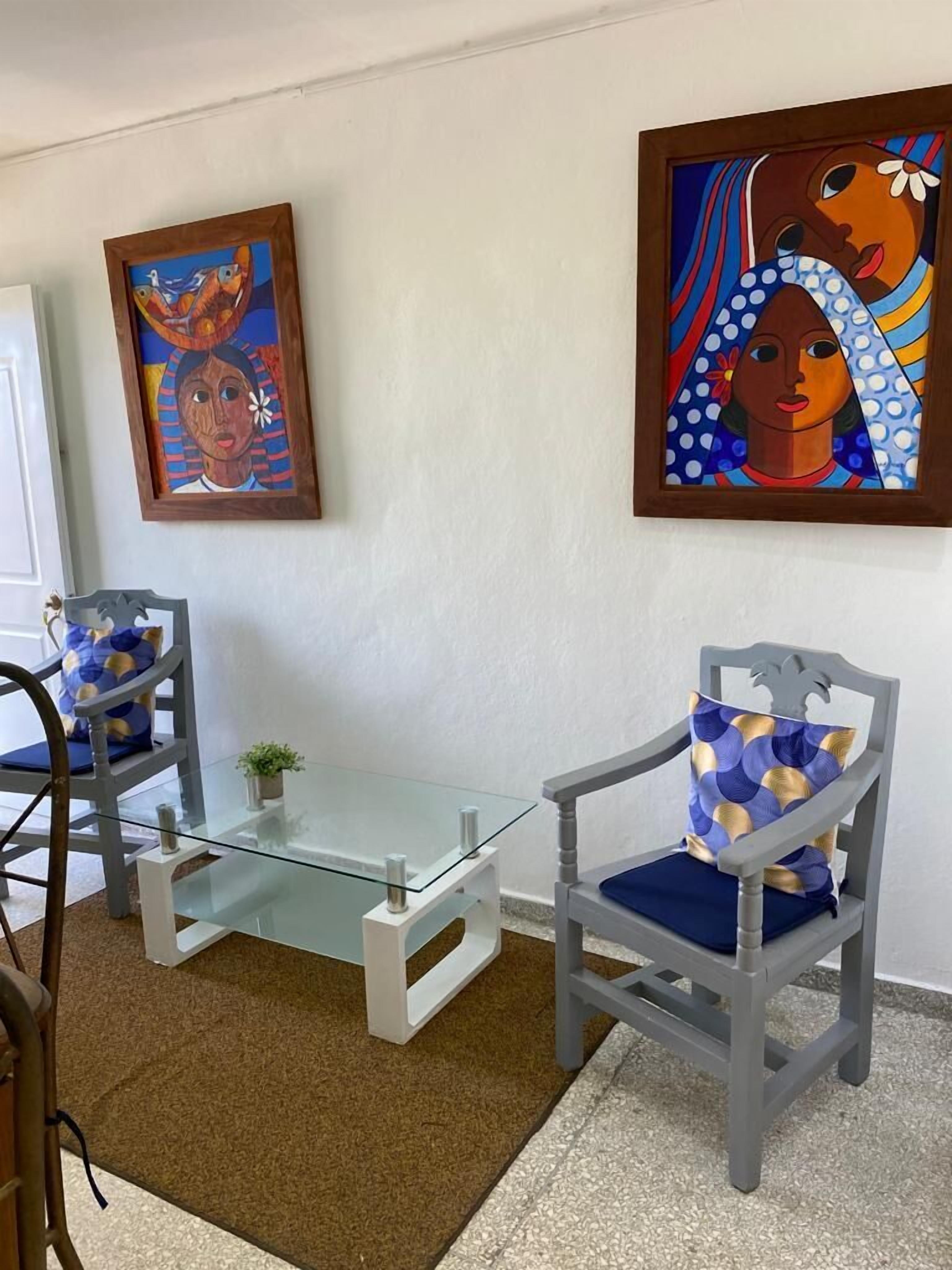 A room with two chairs, a glass table, and two paintings on the wall.