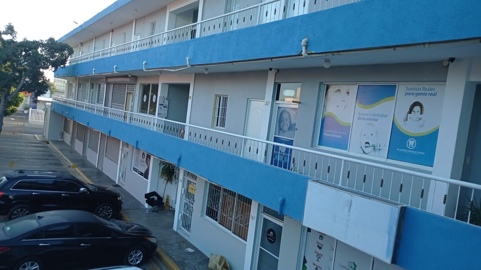 A multi-story building with a blue and white facade, featuring balconies and a parking area with several cars.