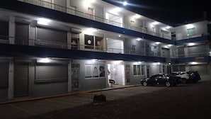 A multi-story building with several closed shutters, a few open doors, and a parking area with cars. - Apartamentos Magallanes Romana (La Romana)