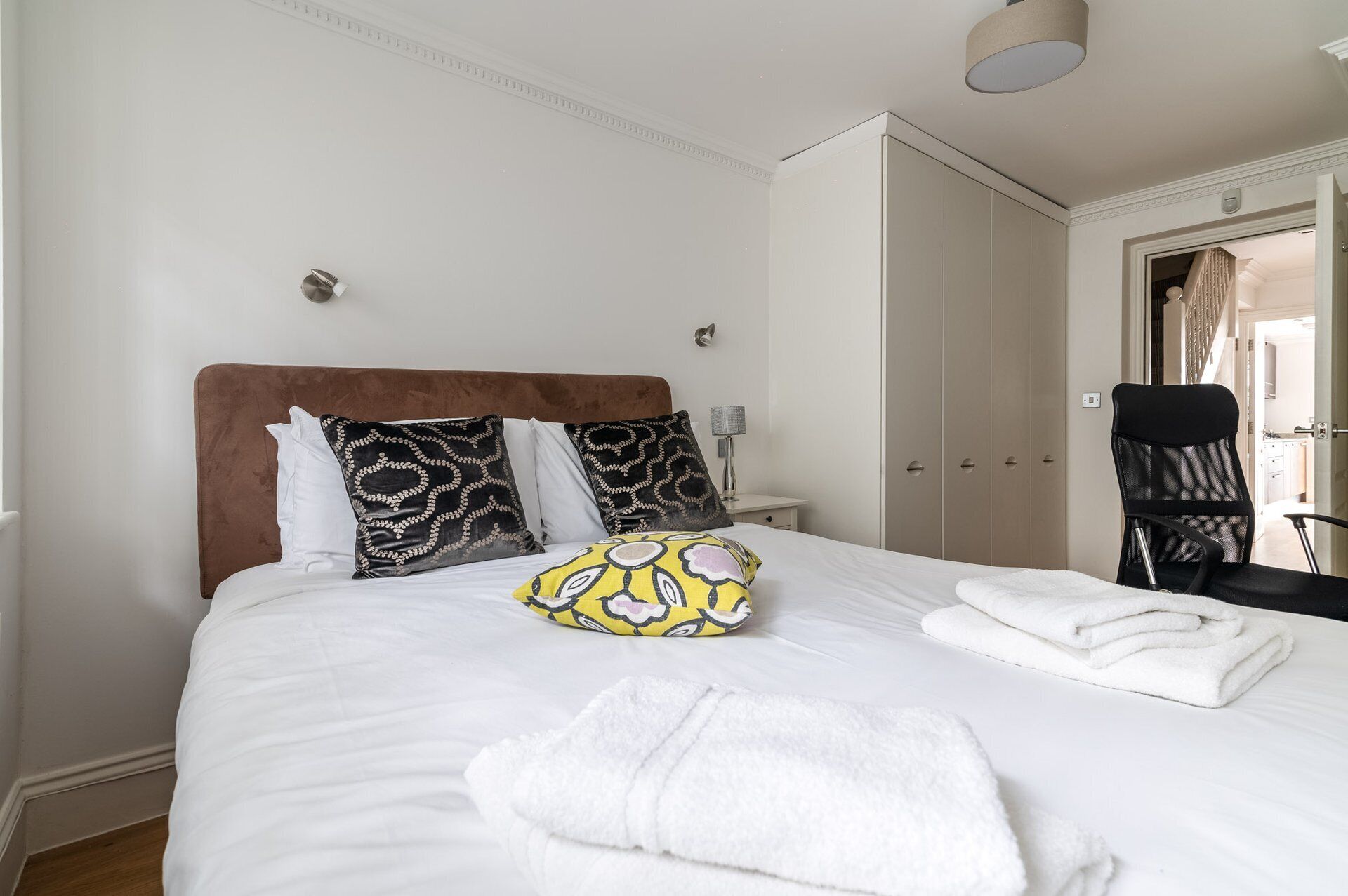 A neatly made bed with white linens, a brown headboard, and decorative pillows. There are white towels on the bed and a chair with a black seat.