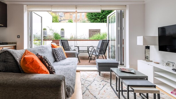 A modern living room with a grey sofa, orange and black cushions, a coffee table, and a TV on a white stand. - Gorgeous Kentish Town Flat (London)