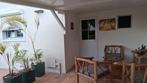 Un patio couvert aménagé avec des meubles en bois, des plantes en pot et un mur blanc agrémenté d’une porte et de fenêtres.