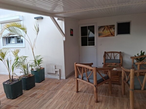 A covered patio area with wooden furniture, potted plants, and a white wall with a door and windows. - Alpinia - Appt avec piscine partagée (Arrondissement of Saint-Pierre)