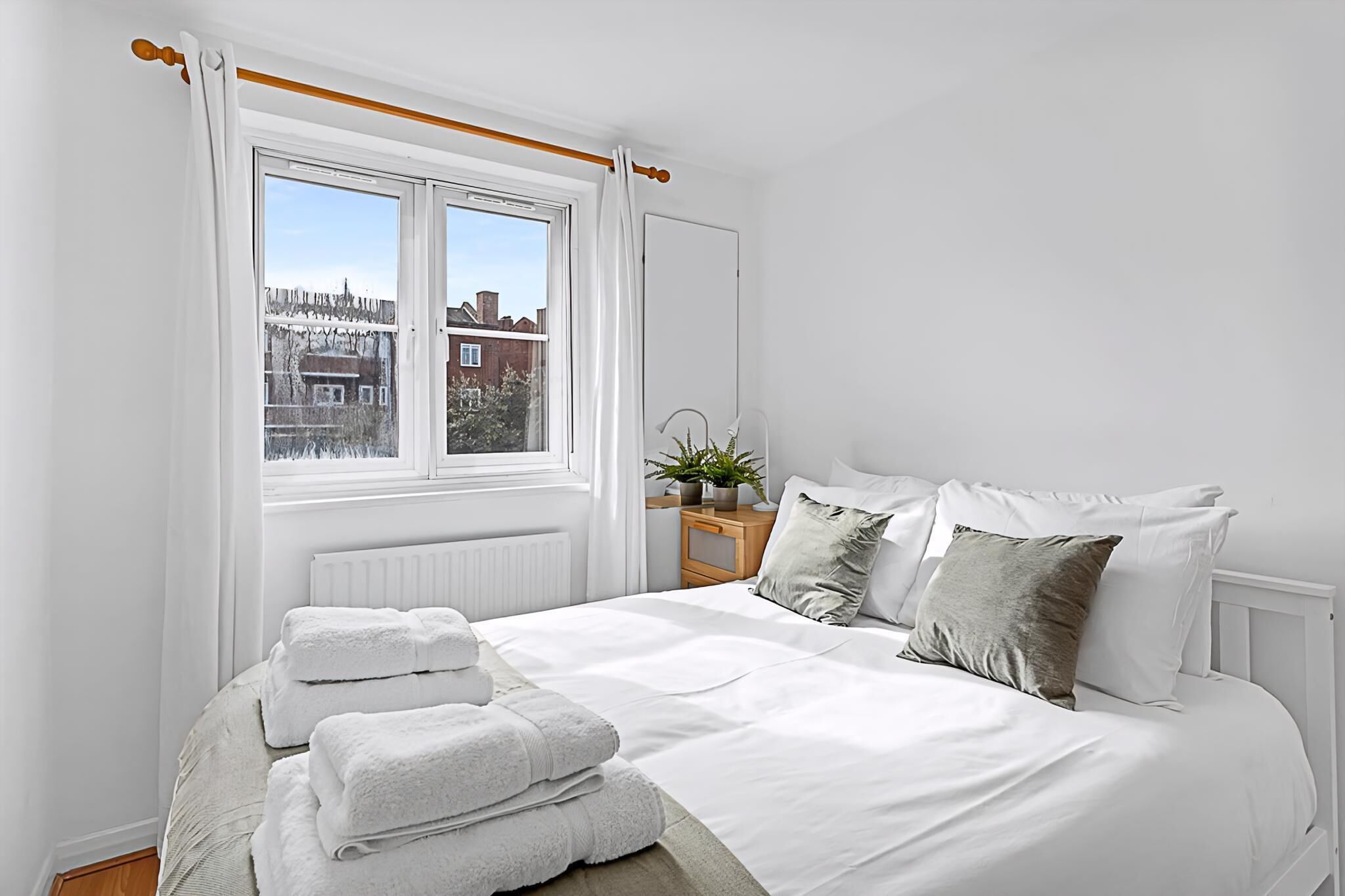 Ein Schlafzimmer mit großem Fenster, einem Bett mit weißer Bettwäsche, einem Nachttisch mit Pflanze und Blick auf Gebäude.
