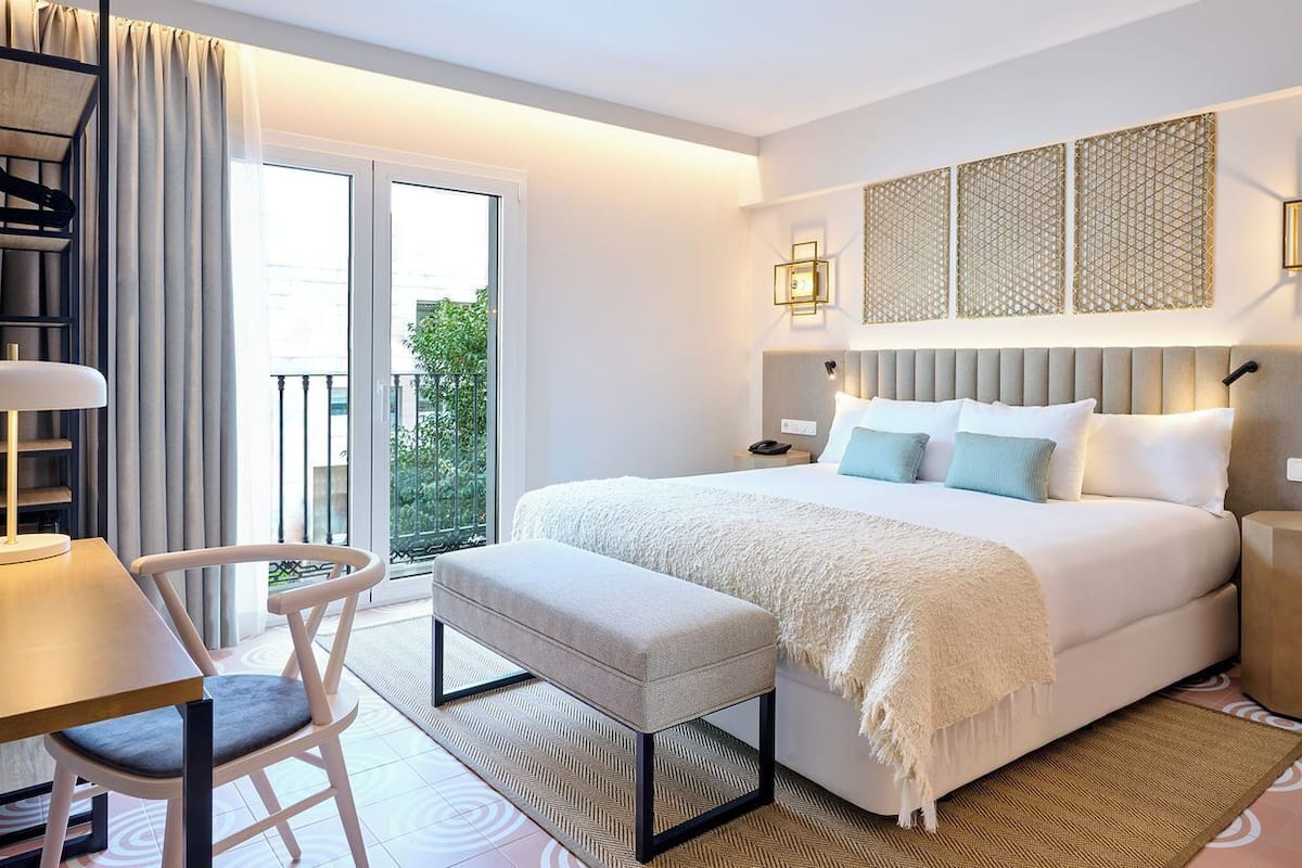 A modern hotel room with a large bed, a desk, and a chair. There is a balcony with a view of greenery outside.