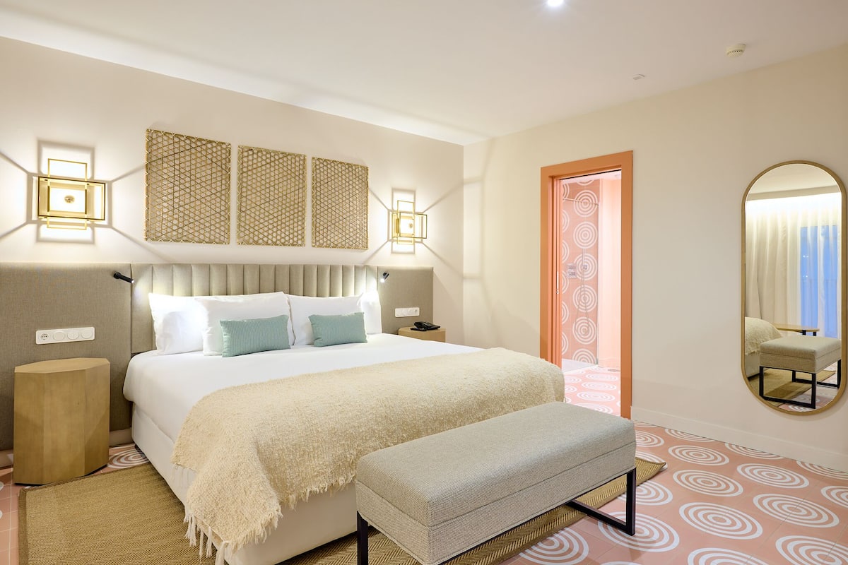 A modern hotel room with a large bed, bedside tables, a bench, a mirror, and patterned floor tiles.