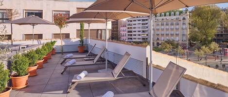 A rooftop terrace with sun loungers, potted plants, and large umbrellas.