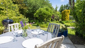 An outdoor dining area with a table set for two, surrounded by lush greenery and a well-maintained garden. - Cosy cottage in a pretty village with a pub at the end of the garden! (Buckland Newton)