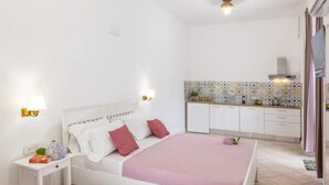 A bedroom with a white bed, a nightstand, and a bedside table. There is a kitchen area with white cabinets and a tiled backsplash. - La Natura x9 (Sorrento)