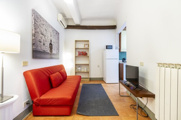Un ambiente compatto con un divano rosso, una TV a schermo piatto, un angolo cottura e un condizionatore d'aria a parete.