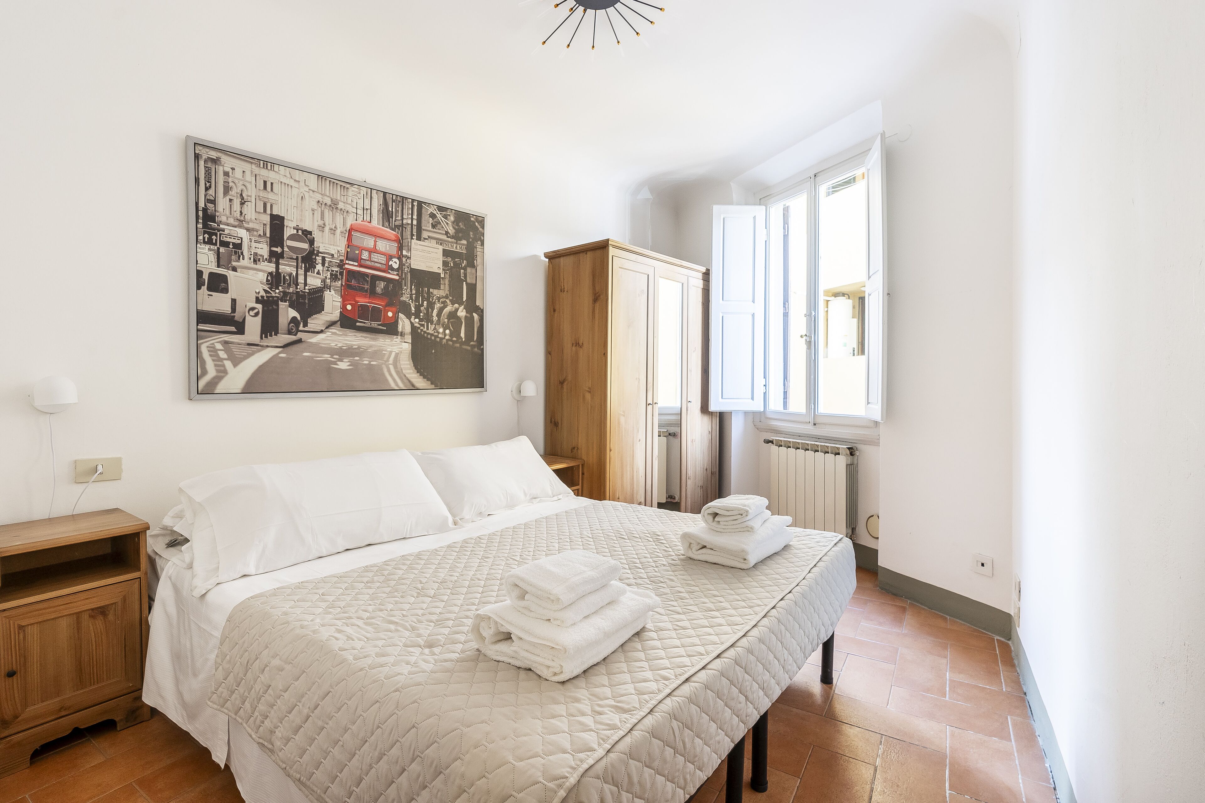 Een net opgemaakt bed met een gebogen wit dekbed, een houten nachtkastje, een houten kledingkast en een groot stadsgezicht schilderij aan de muur.