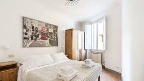 Una cama bien hecha con un edredón blanco acolchado, una mesita de noche de madera, un armario de madera y un cuadro grande con una vista urbana en la pared.