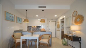 A dining area with a white table and wicker chairs, a kitchen in the background, and a console table with a lamp and decorative items. - Los Flamencos 4004 - Two Bedroom Apartment, Sleeps 5 (Punta del moral)