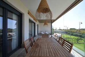 A balcony with a wooden table and chairs, a hanging woven light fixture, and a view of a green landscape.