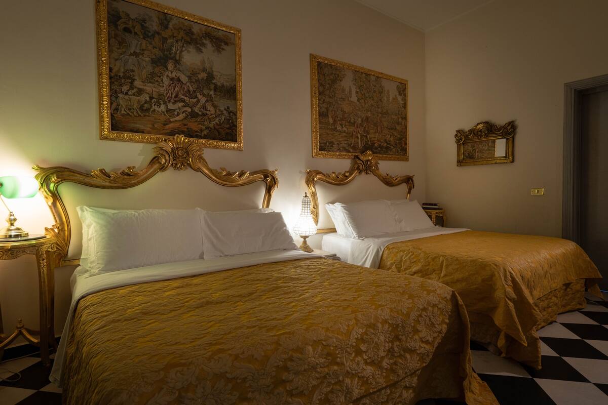 A hotel room with two beds, each with a gold bedspread and white pillows. There are two paintings on the walls, a chandelier, and a small table with a lamp.