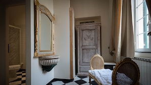 A room with a checkered floor, a bed with a white sheet, a wooden chair, a mirror with a gold frame, and a door with a decorative panel. - San Giorgio Rooms (Genoa)