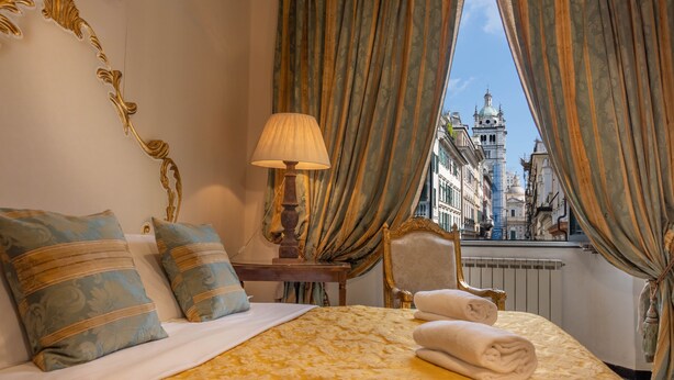 A hotel room with a bed, a lamp, a chair, and a view of a cityscape through curtains. - San Giorgio Rooms (Genoa)
