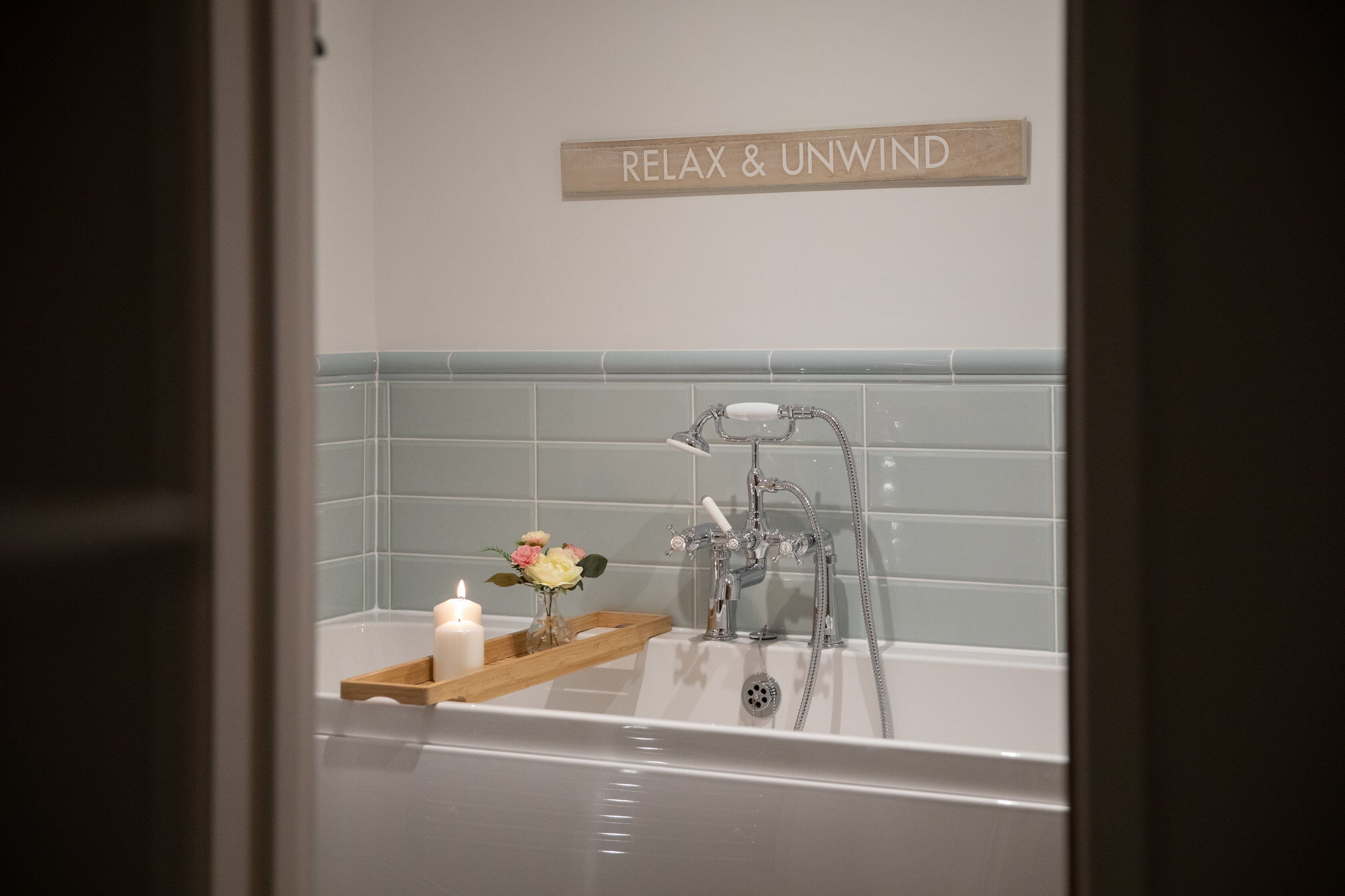 A bathroom with a bathtub, a wooden tray with a candle and flowers, and a sign that reads "RELAX & UNWIND".