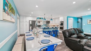 A modern kitchen with white cabinets, a central island, blue bar stools, and a black leather sofa in a living area. - 7BR Pet-Friendly Home w/ EV Charger & Room for 18+ Guests (Bethany Beach)
