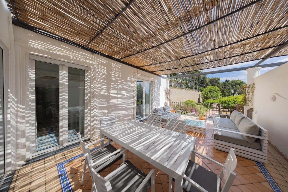 A covered patio area with a table and chairs, a sofa, and a view of the surrounding greenery.