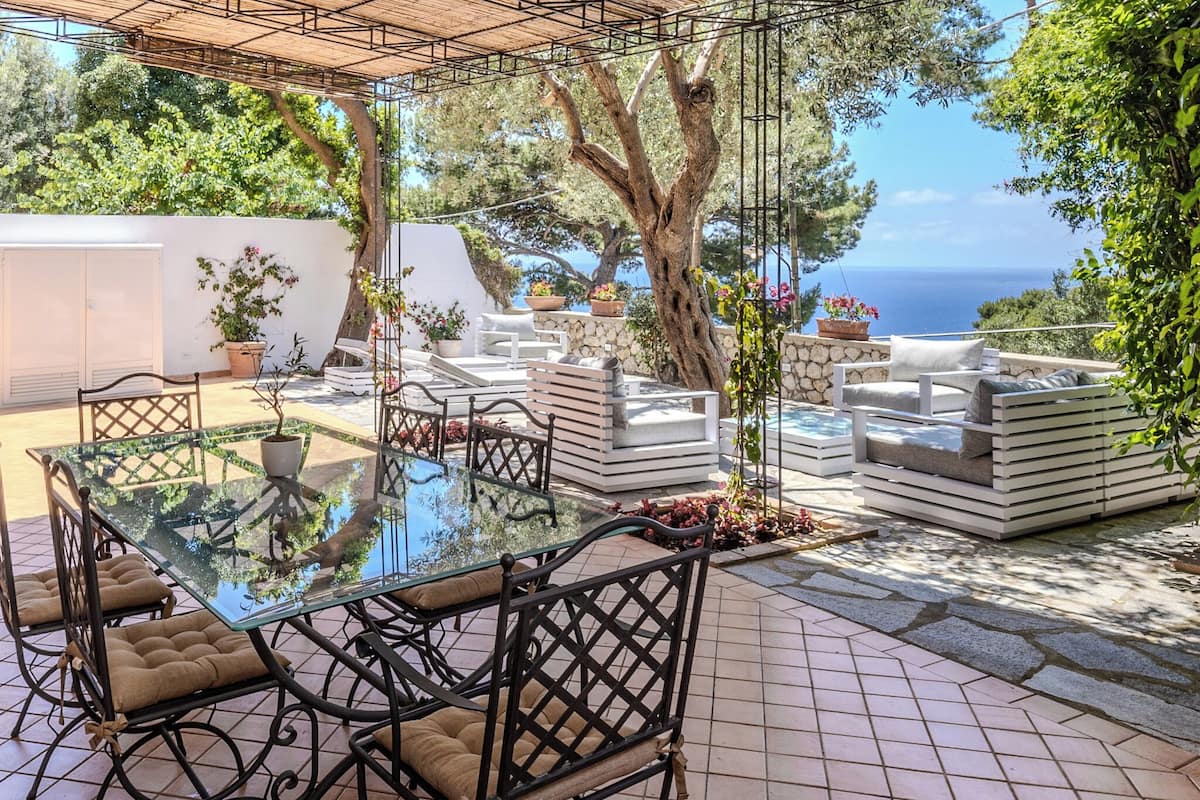 An outdoor patio with a glass table and chairs, surrounded by stone planters and a view of the sea.