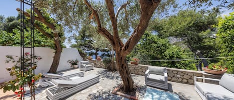 An outdoor patio with stone flooring, white lounge chairs, and a large tree providing shade.