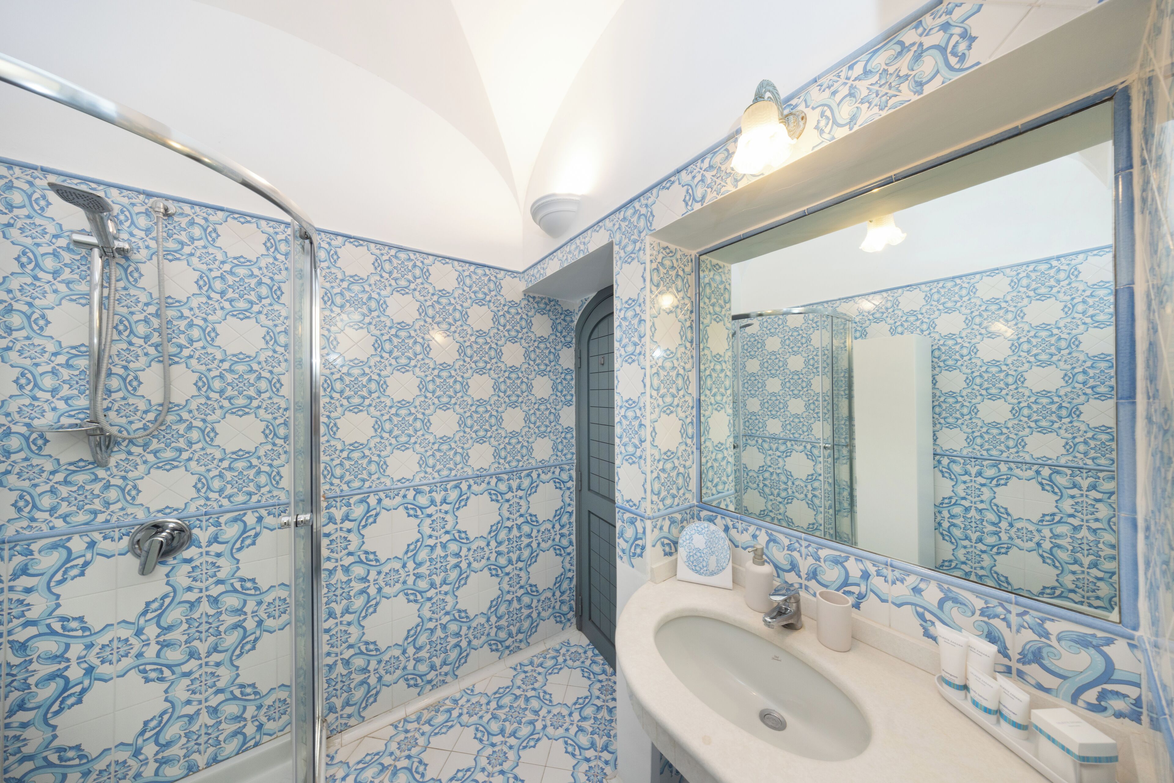 A bathroom with blue patterned tiles, a glass shower enclosure, a sink with toiletries, and a large mirror.