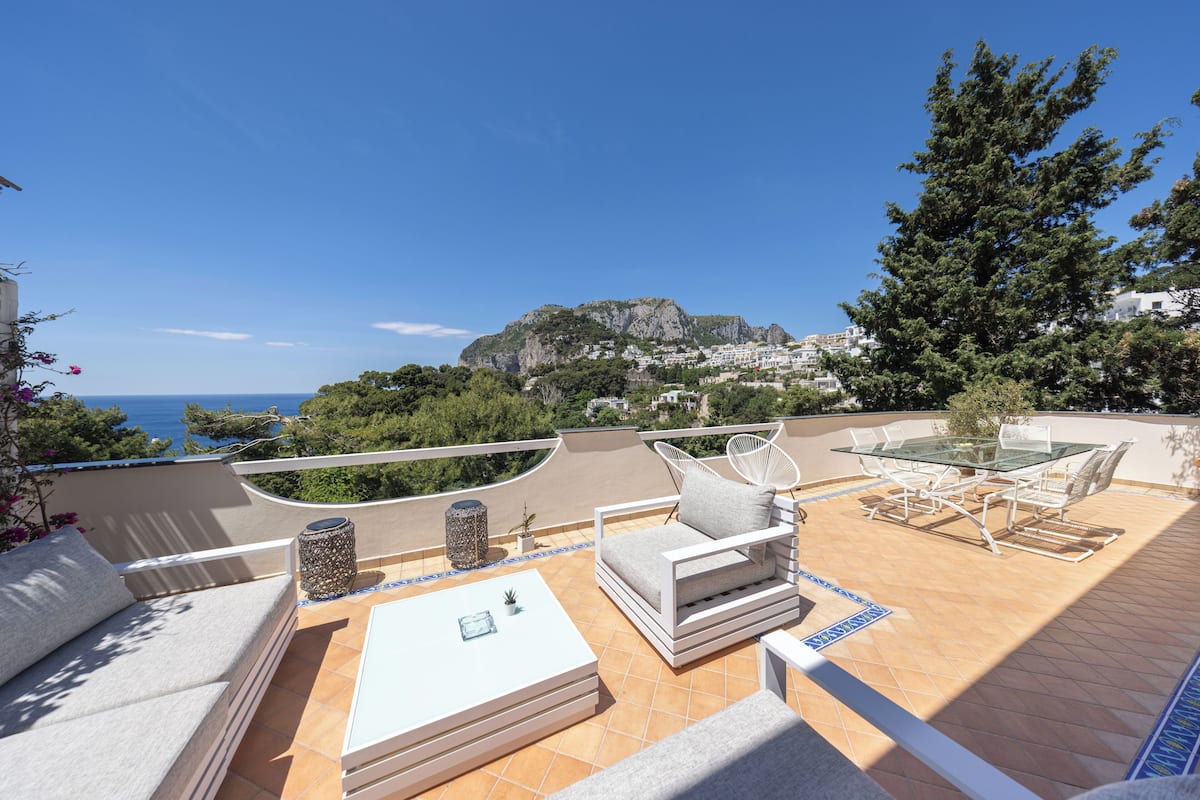 An outdoor patio with a sofa, a table, and chairs, overlooking a scenic view of the sea and a mountain.